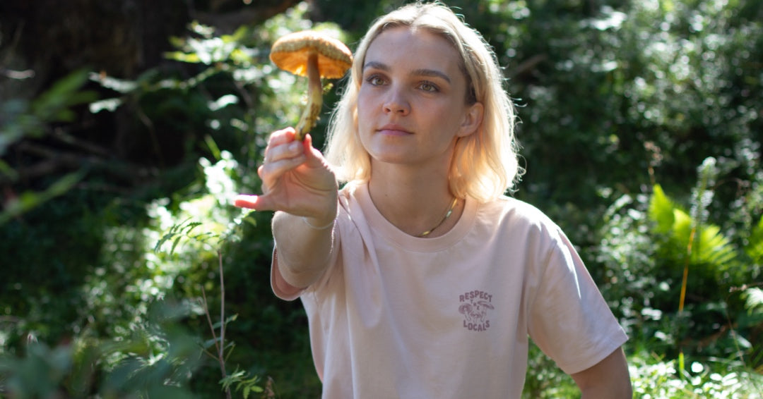Une femme blonde portant un t-shirt NIKIN Mushroom regarde un gros champignon dans sa main tendue.