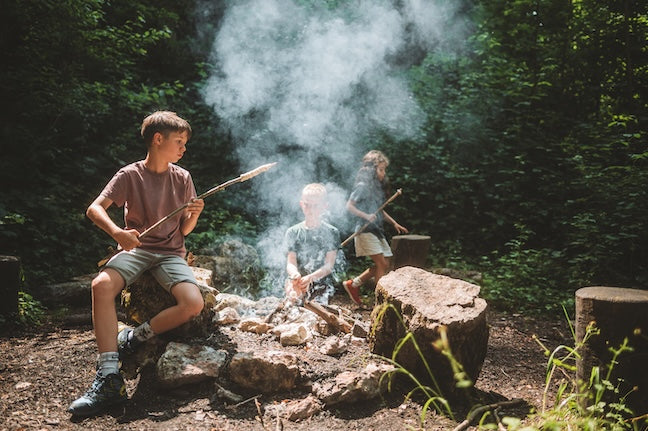 Three children are grilling snake bread over a fire in the forest.