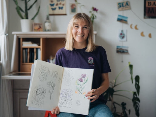 Sophia Hultsch est assise dans son atelier, sourit à la caméra et tient dans sa main un carnet de croquis ouvert avec des dessins floraux. Elle porte un TreeShirt NIKIN violet avec le design "Connected by Nature".