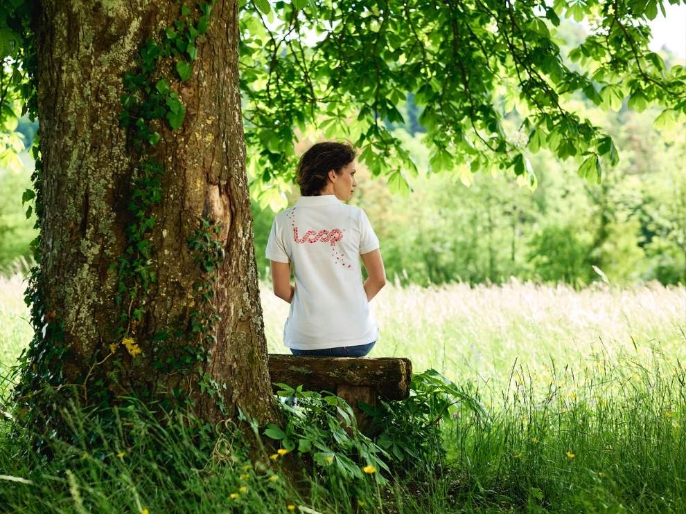 Une femme est assise dehors sous un arbre et porte un shrit blanc avec un logo en boucle qui vient de Pandinavia.